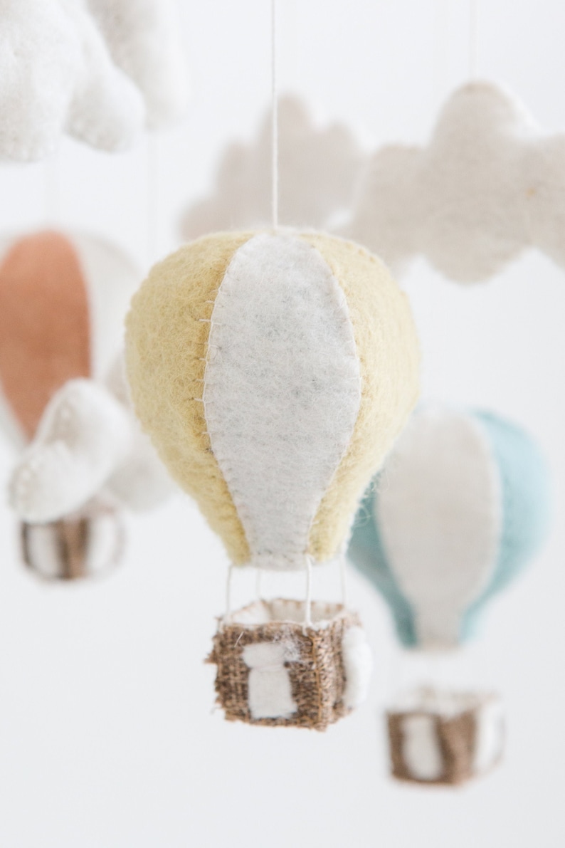 Op de afbeelding: Een handgemaakte mobiel met vilten luchtballonnen in pastelkleuren. De centrale ballon is geel met een witte streep en een geweven mand hangt eronder. Andere ballonnen zijn perzik en blauw, met witte vilten wolken.