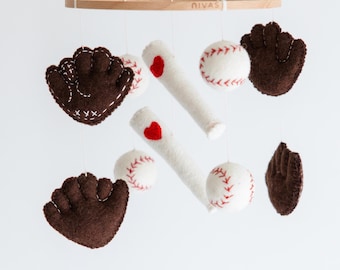 May include: A baseball-themed baby mobile featuring felt baseballs with red stitching, white felt bats with red hearts, and brown felt baseball gloves. The mobile hangs from a wooden ring.