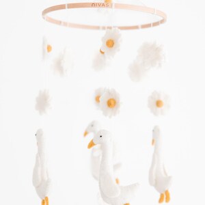 May include: A white felt baby mobile with a wooden ring, featuring white felt geese, white felt clouds, and yellow-centered white felt daisies. The ring has the word "Nivas" printed on it.
