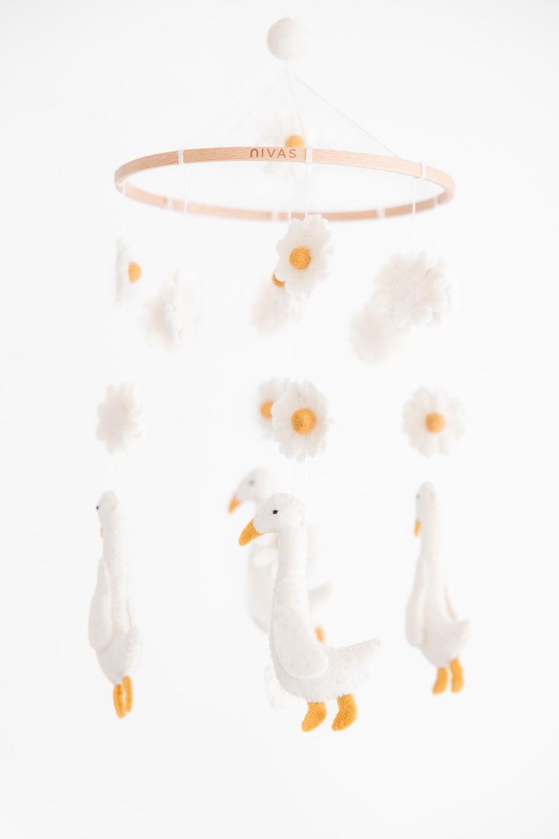 May include: A white felt baby mobile with a wooden ring, featuring white felt geese, white felt clouds, and yellow-centered white felt daisies. The ring has the word "Nivas" printed on it.