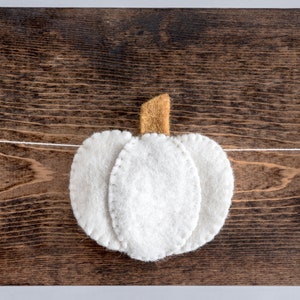 May include: A white felt garland with three pumpkins, each with a brown felt stem. The garland is strung on a white thread and is hanging on a brown wooden surface.