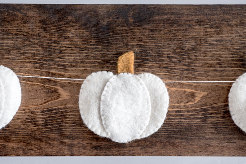 May include: A white felt garland with three pumpkins, each with a brown felt stem. The garland is strung on a white thread and is hanging on a brown wooden surface.