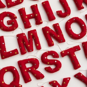 May include: A collection of red felt alphabet letters, each featuring small gold star-shaped embellishments and silver sequins. Every letter includes a thin white string loop attached to the top, designed for hanging as individual ornaments.