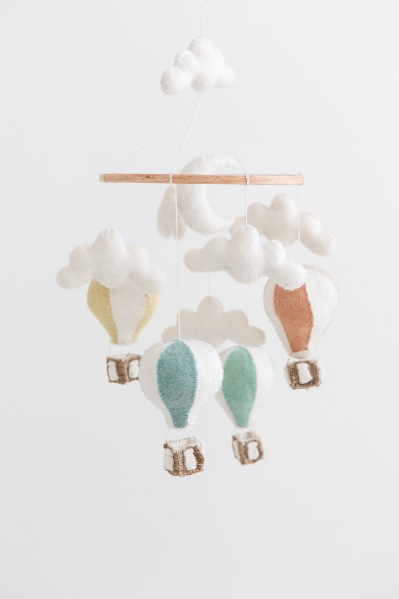 Op de afbeelding: Een baby mobile met vilten luchtballonnen in pastelkleuren, wolken en een maan. De ballonnen hebben witte, gele, blauwgroene en bruine accenten, hangend aan een houten ring.