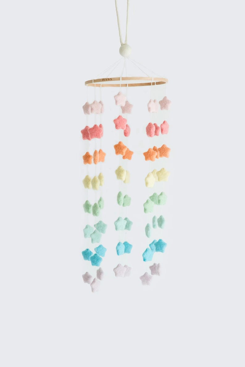 May include: A baby mobile featuring a wooden ring with hanging felt stars in a rainbow of colors. The stars are arranged in vertical strands, transitioning from pink to blue. The mobile is suspended by a white cord.
