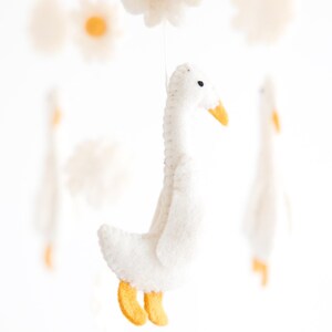 May include: A white felt goose mobile hanging from a white string with yellow felt feet and beak. The goose is surrounded by white felt daisies with yellow centers.