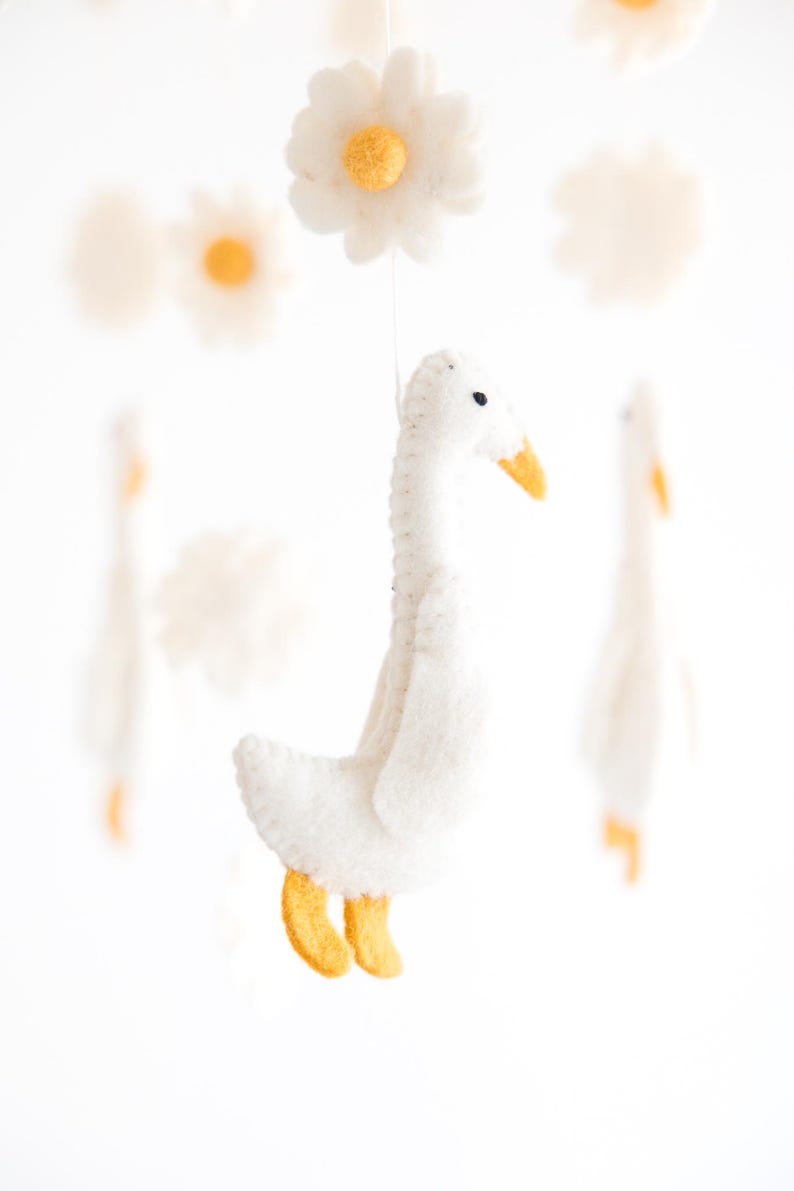 May include: A white felt goose mobile hanging from a white string with yellow felt feet and beak. The goose is surrounded by white felt daisies with yellow centers.