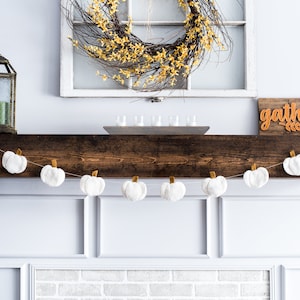 May include: A white brick fireplace with a white fluffy pumpkin garland hanging above it. A wreath with yellow flowers is hanging on a white window above the fireplace. The word "gather" is written on a wooden sign above the fireplace.