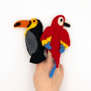 May include: Two felt finger puppets, one a red and yellow parrot and the other a black and yellow toucan, are worn on a person's fingers.