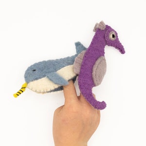 May include: A purple felt seahorse finger puppet with a gray felt whale finger puppet on its back. Both puppets have embroidered details.