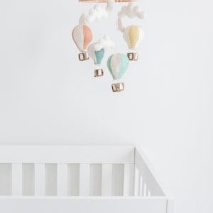 Op de afbeelding: Een baby mobile met kleurrijke luchtballonnen in pastel tinten perzik, geel en teal, hangend aan een houten ring. Witte wolken accenten en een wit babybedje zijn ook zichtbaar.
