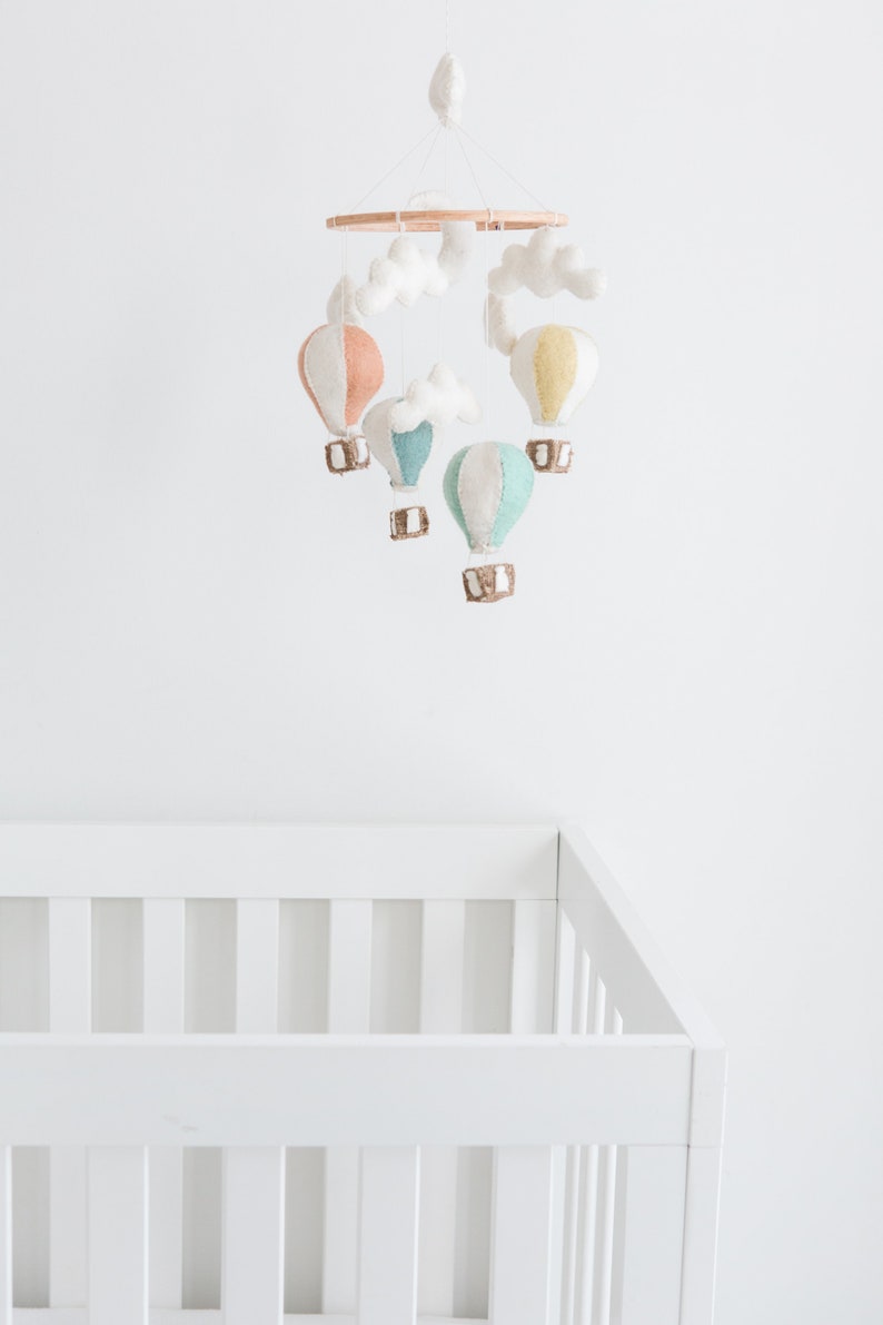 Op de afbeelding: Een baby mobile met kleurrijke luchtballonnen in pastel tinten perzik, geel en teal, hangend aan een houten ring. Witte wolken accenten en een wit babybedje zijn ook zichtbaar.