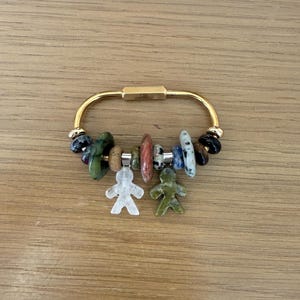 May include: A gold-colored metal keychain with a variety of colorful beads and two small, carved human figures. The beads include green, red, blue, and black stones. The keychain is on a wooden surface.
