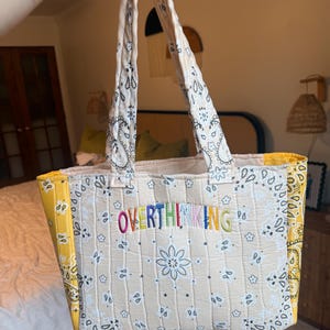 Puede incluir: Un bolso tote acolchado con estampado de bandana en color crema, motivos florales negros y paneles laterales en amarillo brillante. La palabra OVERTHINKING est&aacute; bordada en hilo multicolor en el centro, con dos asas de tela a juego.
