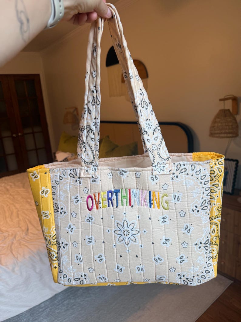 Puede incluir: Un bolso tote acolchado con estampado de bandana en color crema, motivos florales negros y paneles laterales en amarillo brillante. La palabra OVERTHINKING est&aacute; bordada en hilo multicolor en el centro, con dos asas de tela a juego.