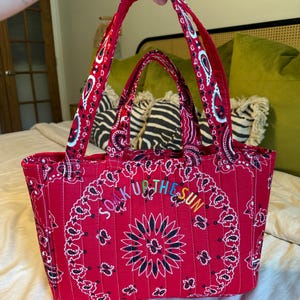 Puede incluir: Un bolso tote rojo acolchado con un patr&oacute;n tradicional de bandana de cachemira en blanco y negro. El bolso tiene dos asas de tela y el texto bordado "SOAK UP THE SUN" en letras multicolores arqueadas en el panel central delantero.