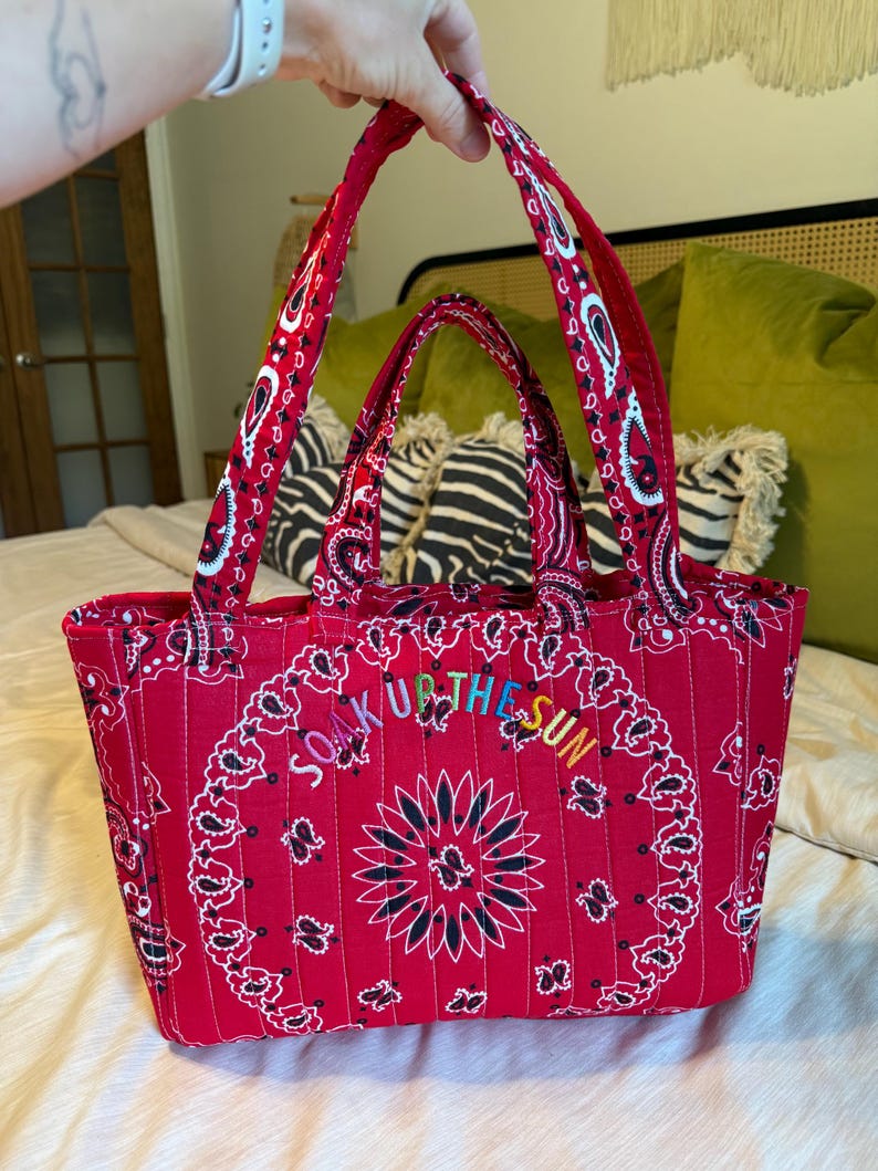 Puede incluir: Un bolso tote rojo acolchado con un patr&oacute;n tradicional de bandana de cachemira en blanco y negro. El bolso tiene dos asas de tela y el texto bordado "SOAK UP THE SUN" en letras multicolores arqueadas en el panel central delantero.