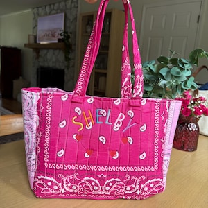 Puede incluir: Un bolso tote acolchado de color rosa vibrante con un estampado de cachemira blanco estilo bandana. El frente muestra el nombre "SHELBY" bordado en letras multicolores sobre tres peque&ntilde;as aplicaciones rojas en forma de coraz&oacute;n. Tiene dos asas a juego.