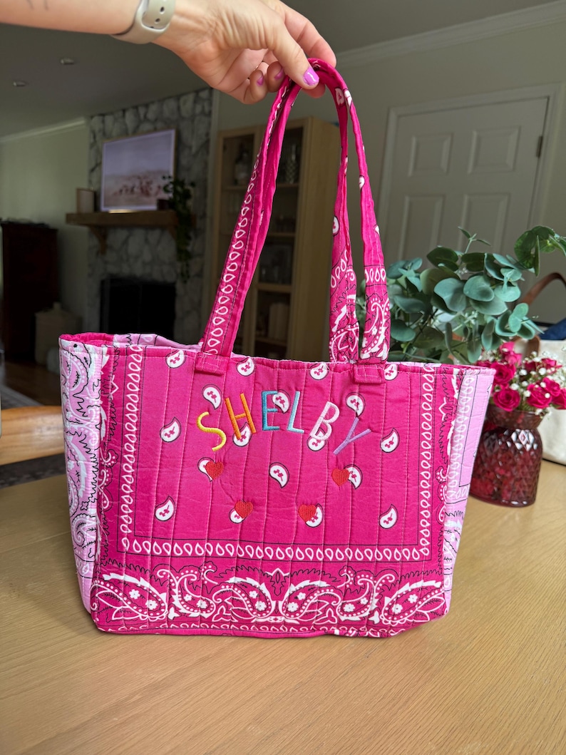 Puede incluir: Un bolso tote acolchado de color rosa vibrante con un estampado de cachemira blanco estilo bandana. El frente muestra el nombre "SHELBY" bordado en letras multicolores sobre tres peque&ntilde;as aplicaciones rojas en forma de coraz&oacute;n. Tiene dos asas a juego.