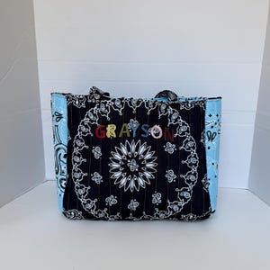 Puede incluir: Un bolso tote acolchado con estampado de bandana en blanco y negro en la parte frontal y estampado de bandana azul claro en los laterales. El nombre GRAYSON est&aacute; bordado con letras multicolores en el centro del panel de tela negra.