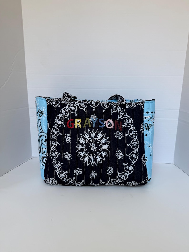 Puede incluir: Un bolso tote acolchado con estampado de bandana en blanco y negro en la parte frontal y estampado de bandana azul claro en los laterales. El nombre GRAYSON est&aacute; bordado con letras multicolores en el centro del panel de tela negra.