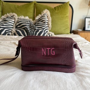 May include: A burgundy faux-leather cosmetic bag with a crocodile skin texture. The bag has a zipper closure and a small handle on each side. The initials "NTG" are embroidered in pink on the front.