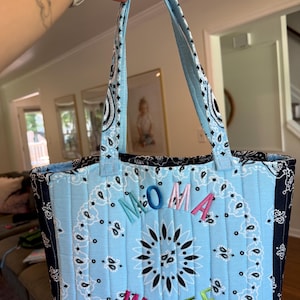 Puede incluir: Un bolso tote acolchado con un patr&oacute;n de bandana de cachemira en azul claro y negro. El bolso tiene dos asas a juego y el texto bordado MOMA WOLFE en letras multicolores en el centro de la parte frontal de la tela acolchada.