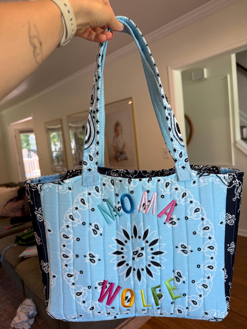 Puede incluir: Un bolso tote acolchado con un patr&oacute;n de bandana de cachemira en azul claro y negro. El bolso tiene dos asas a juego y el texto bordado MOMA WOLFE en letras multicolores en el centro de la parte frontal de la tela acolchada.