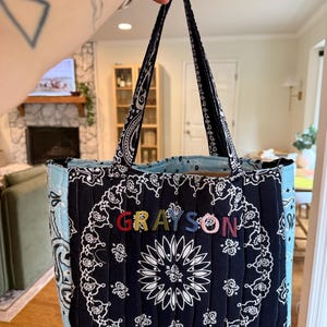 Puede incluir: Un bolso tote acolchado con estampado de bandana en blanco y negro y paneles laterales en azul claro. La parte frontal del bolso muestra el nombre GRAYSON en letras bordadas de varios colores. Tiene dos asas de tela a juego para llevarlo.