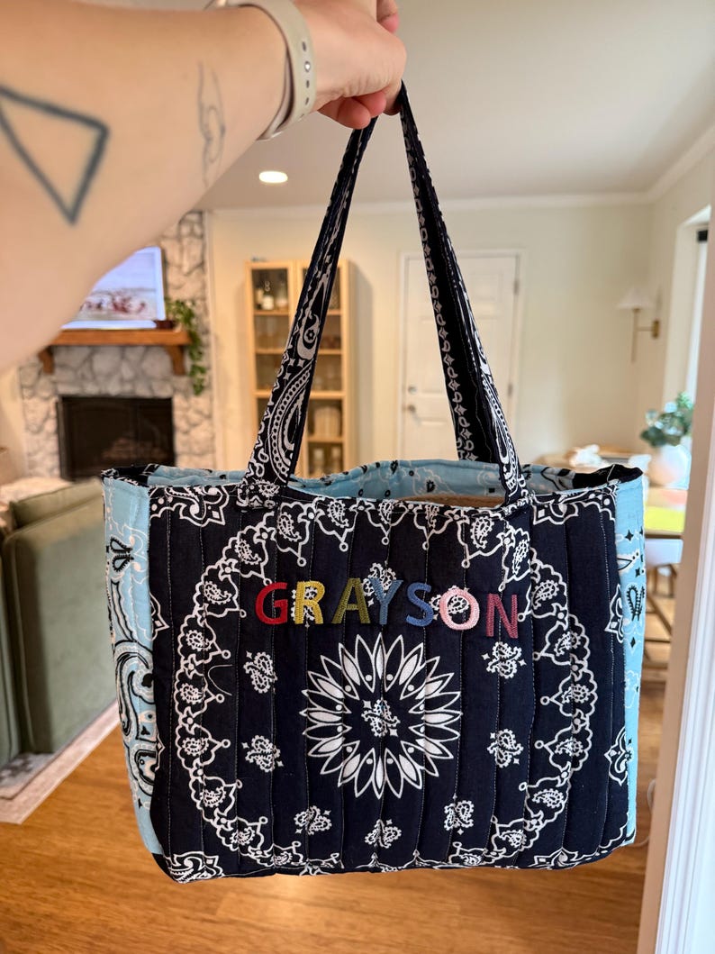 Puede incluir: Un bolso tote acolchado con estampado de bandana en blanco y negro y paneles laterales en azul claro. La parte frontal del bolso muestra el nombre GRAYSON en letras bordadas de varios colores. Tiene dos asas de tela a juego para llevarlo.