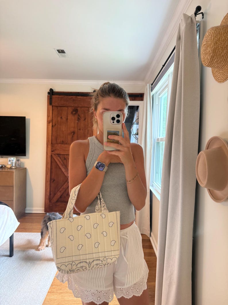 Puede incluir: Un bolso de mano peque&ntilde;o, rectangular y acolchado, confeccionado en tela de color crema con un estampado de pa&ntilde;uelo de cachemira en negro. El bolso cuenta con dos asas cortas de tela y una forma estructurada con costuras visibles en el exterior.