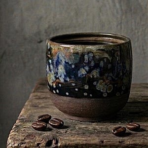 May include: A ceramic coffee mug with a dark brown base and a speckled blue, brown, and white upper section. Several coffee beans are scattered on the rough wooden surface. The mug sits on a weathered wooden table.