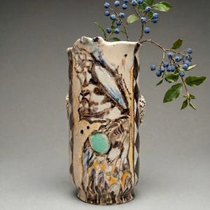 May include: A decorative ceramic vase with a textured surface and an artistic design. The vase is predominantly brown and beige, with blue and turquoise accents. It contains a sprig of blueberries with green leaves, creating a natural contrast.