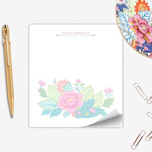 Könnte beinhalten: Ein weißer Notizblock mit floralem Design in Pastellfarben, ein goldener Stift und goldene Büroklammern. Der Notizblock trägt oben den Namen "RACHEL BIRNBAUM". Im Hintergrund befindet sich ein dekorativer Teller mit floralem Design.