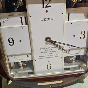 May include: A Seiko wall clock with a brown wooden frame and a white face. The clock features black numerals, hands, and the text "SEIKO". The clock has a glass cover and decorative elements.