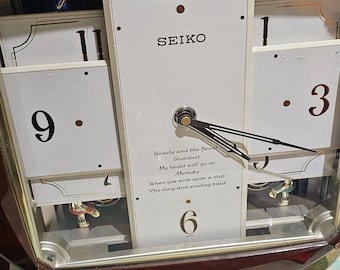 May include: A Seiko wall clock with a brown wooden frame and a white face. The clock features black numerals, hands, and the text "SEIKO". The clock has a glass cover and decorative elements.