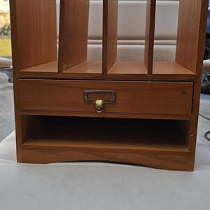 May include: A wooden desk organizer with multiple vertical slots and a drawer. The organizer is made of light brown wood and features a brass-colored drawer pull. The design is suitable for office or home use.