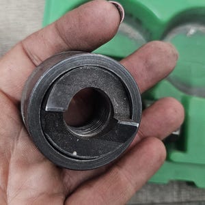 Puede incluir: Una herramienta de metal gris oscuro con un dise&ntilde;o circular y un interior roscado. La herramienta se sostiene en una mano, con una caja de pl&aacute;stico verde visible en el fondo. La herramienta parece ser parte de un conjunto m&aacute;s grande.