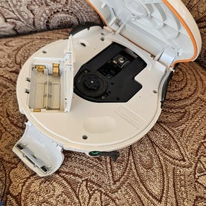 Op de afbeelding: Een witte draagbare cd-speler met een open deksel en een blootgesteld batterijcompartiment. Het apparaat heeft een cirkelvormig ontwerp met een oranje rand, een zwart centraal schijfmechanisme en een klein waarschuwingslabel.