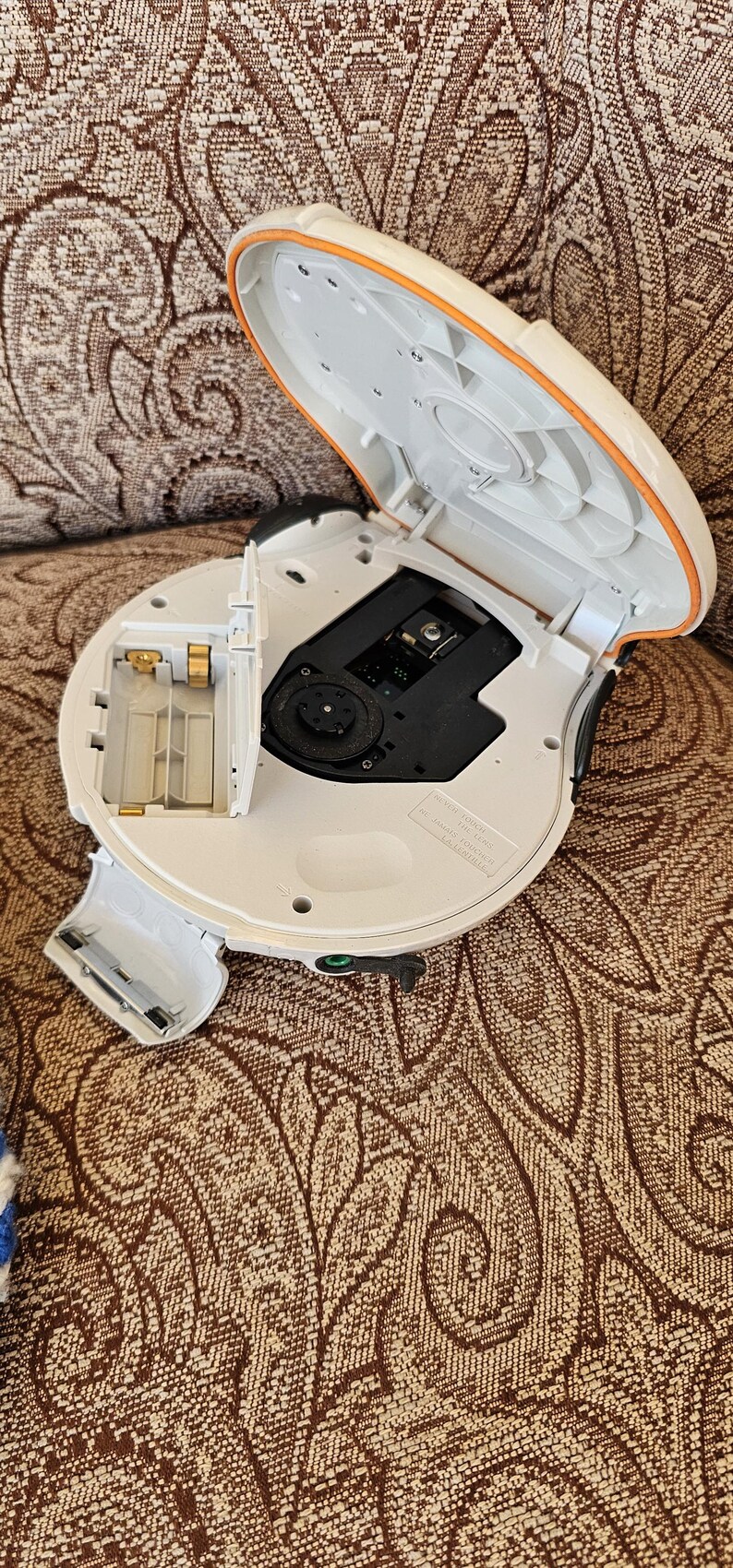 Op de afbeelding: Een witte draagbare cd-speler met een open deksel en een blootgesteld batterijcompartiment. Het apparaat heeft een cirkelvormig ontwerp met een oranje rand, een zwart centraal schijfmechanisme en een klein waarschuwingslabel.