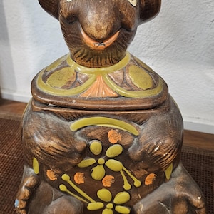 May include: A brown ceramic cookie jar shaped like a turtle with a hat. The turtle has a smiling face with brown eyes and a yellow nose. The jar is decorated with green, orange, and yellow flowers.