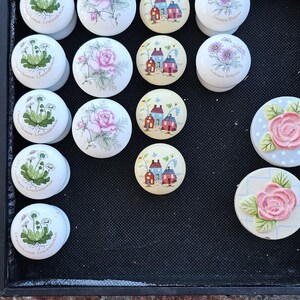 May include: A collection of decorative ceramic knobs with floral and house designs. The knobs feature pink roses, purple flowers, and small houses with red roofs. Some knobs have the text "Chrysanthemum Leucanthemum" and "Gazania Ringens".