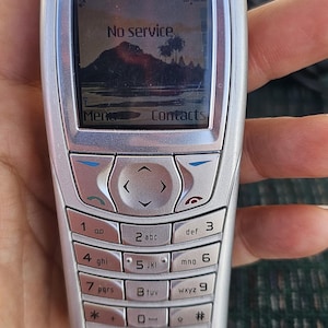 May include: A silver Nokia cell phone with a black screen that reads "No service". The phone has a keypad with numbers and letters, and a small AT&T logo on the bottom.
