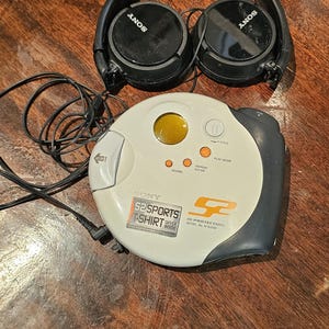 Op de afbeelding: Een witte en donkergrijze Sony S2 Sports cd-speler met een rond ontwerp, voorzien van een geel venster, bedieningsknoppen en G-Protection-technologie. Het apparaat bevat een zwarte bedrade hoofdtelefoon met het Sony-logo op de oorschelpen.