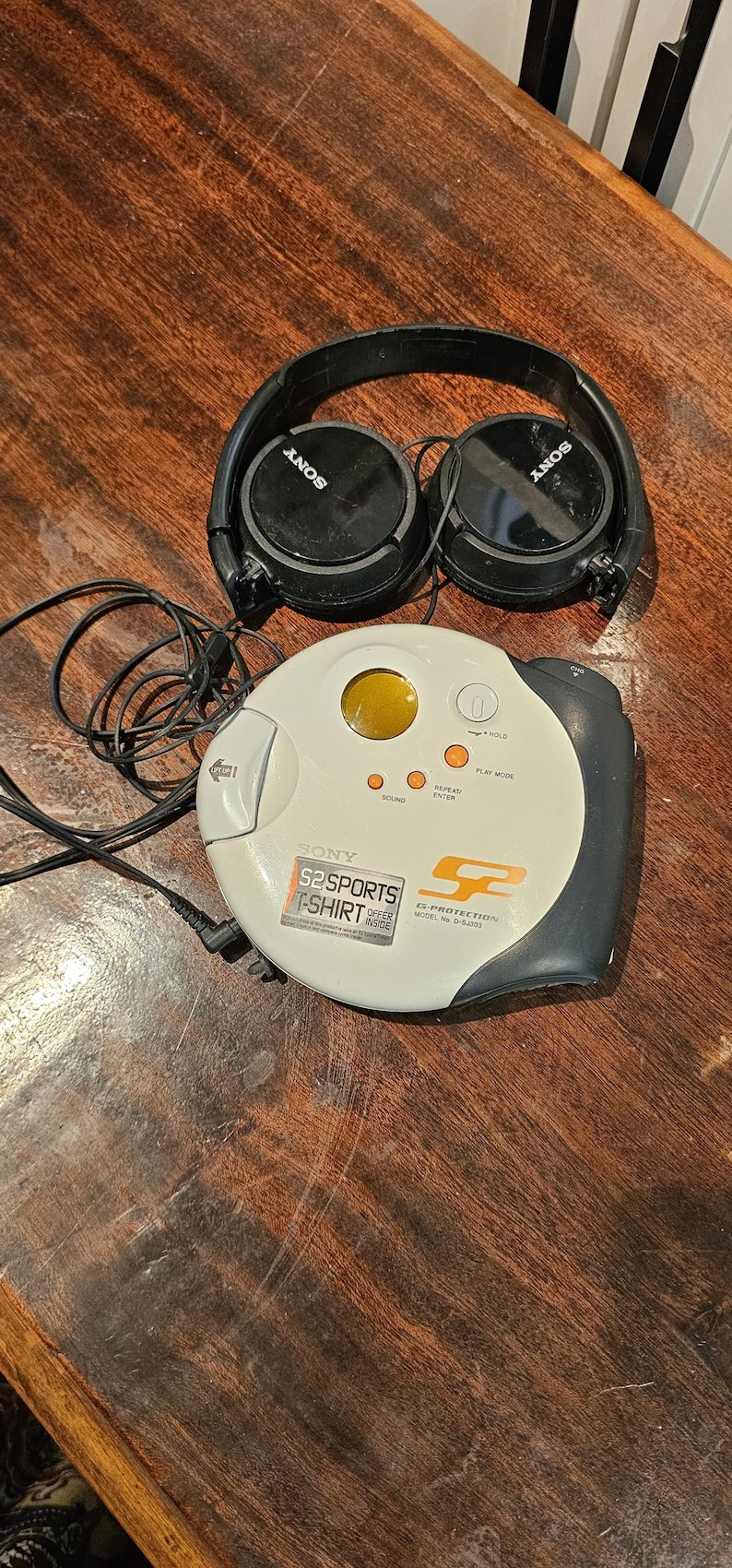 Op de afbeelding: Een witte en donkergrijze Sony S2 Sports cd-speler met een rond ontwerp, voorzien van een geel venster, bedieningsknoppen en G-Protection-technologie. Het apparaat bevat een zwarte bedrade hoofdtelefoon met het Sony-logo op de oorschelpen.