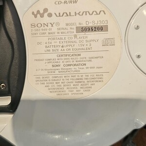Op de afbeelding: Een witte Sony Walkman draagbare cd-speler, model D-SJ303, met een cirkelvormig ontwerp. Het label aan de achterkant toont technische specificaties, waaronder DC 4.5V, batterijvereisten van 1.5V x 2 LR6 AA en serienummer 5098200.