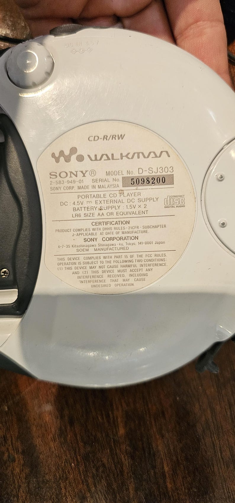 Op de afbeelding: Een witte Sony Walkman draagbare cd-speler, model D-SJ303, met een cirkelvormig ontwerp. Het label aan de achterkant toont technische specificaties, waaronder DC 4.5V, batterijvereisten van 1.5V x 2 LR6 AA en serienummer 5098200.