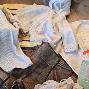 May include: A pile of baby clothes including a white hooded sweater, a pair of light blue knitted pants, a white bib with a cartoon baby on it, and a light blue knitted cardigan. The bib has the text 