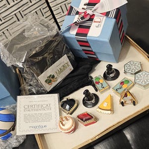 May include: A collection of small, colorful, and decorative trinkets, including a blue and white striped ball, a small cake, a red jewel, a slice of pie, and a few other trinkets. The trinkets are arranged on a white surface with a gold trim. The trinkets are surrounded by a variety of other items, including a blue box with the word "artfoam" on it, a black box with the word "lucky" on it, and a certificate of authenticity.