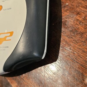 Op de afbeelding: Een draagbaar elektronisch apparaat met een mat donkergrijs zijpaneel en een wit bovenoppervlak. De bovenkant is voorzien van een ronde knop met het label HOLD, een PLAY MODE-knop en oranje tekst met het woord PROTECTION.